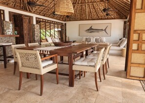 A dining area with a wooden table and wicker chairs, a thatched roof, and a large fish painting on the wall.