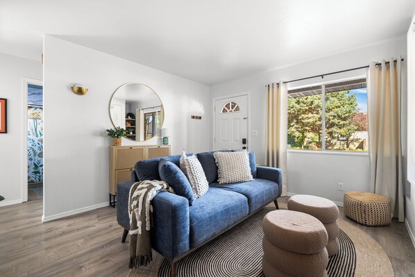 Una sala de estar moderna con un sofá azul, un espejo redondo y vista a árboles por una ventana.