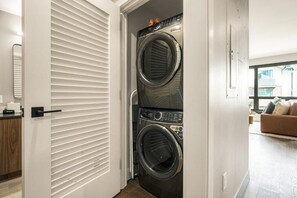 A laundry room with two stacked front-loading washing machines, a door with horizontal blinds, and a view of a living area with a sofa. - Kindred Resort Luxury Ski In, Ski Out 1 Bedroom Residence at the River Run Gondola (Keystone)