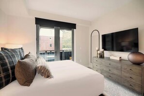 Een slaapkamer met een bed, een dressoir, een flatscreen televisie en een raam met uitzicht op een gebouw en bomen.