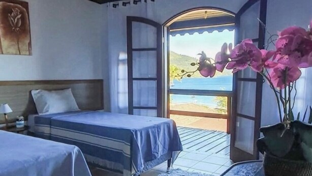 A room with two beds, a wooden ceiling, a view of the sea through an open door, and a vase with pink flowers on a table. - Pousada Três Coqueiros (Angra dos Reis)