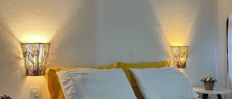 A bedroom with a bed, two bedside lamps, a small table with a plant, and a painting on the wall.