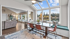 A spacious living room with a glass dining table, comfortable seating, a ceiling fan, and large windows offering a view of a lake and trees.