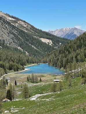 Paisaje montañoso con un lago, colinas boscosas y una cabaña pequeña.