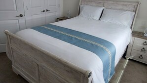 Un dormitorio con cama de madera, ropa de cama blanca, un detalle azul en la colcha, una mesita de noche con una lámpara y una puerta cerrada.
