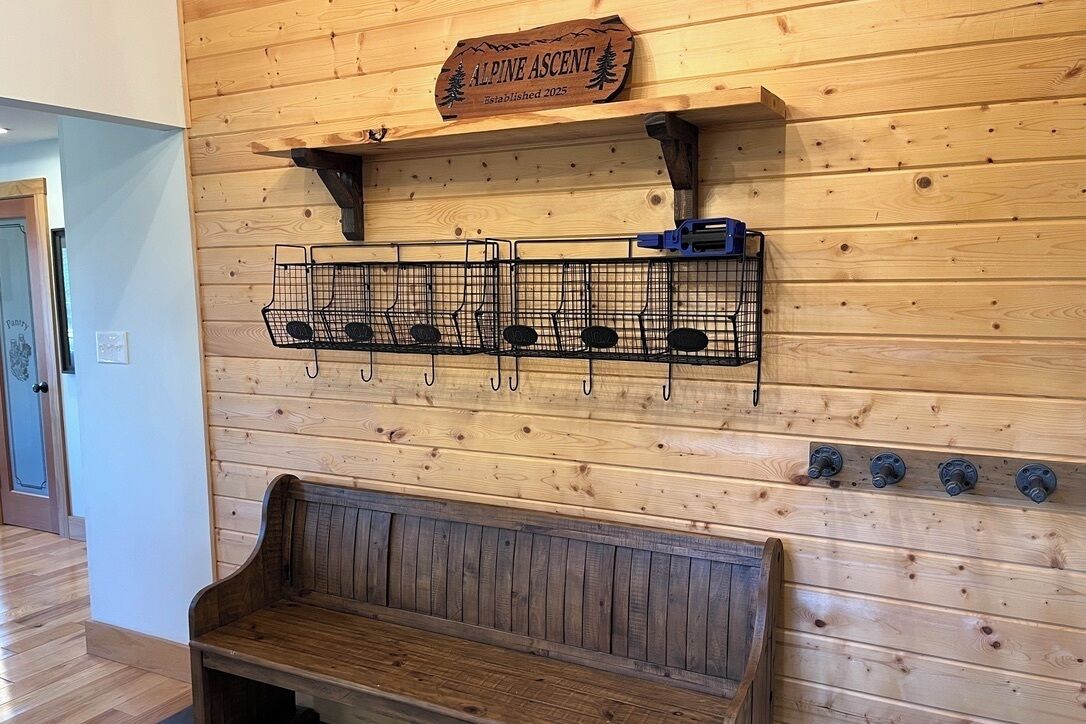 A wooden wall with a sign reading 'Alpine Ascent Established 2025,' a metal rack with hooks, and a wooden bench.
