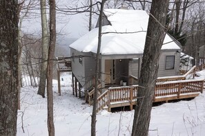 Une cabane dotée d’une terrasse en bois et d’un toit enneigé, entourée d’arbres.
