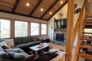 Un salon chaleureux avec une cheminée en pierre, un escalier en bois et une grande fenêtre.
