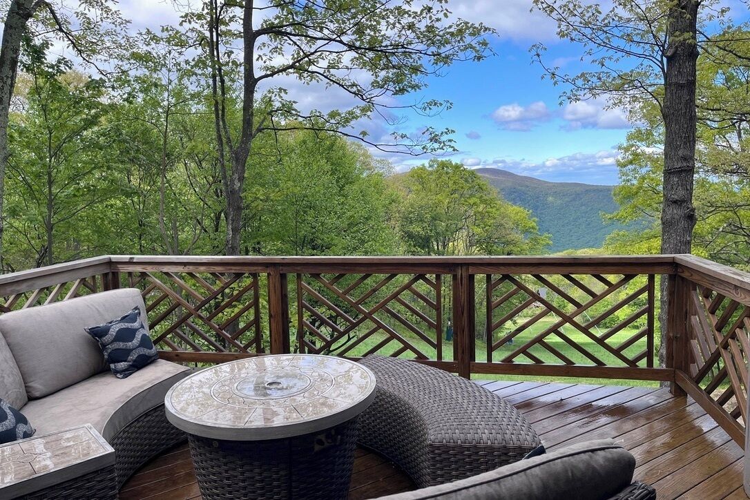 A deck with a round table and a sofa, surrounded by a wooden railing, overlooking a forested landscape.