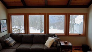 A room with a wooden ceiling, a sofa with cushions, a small table, and three windows with a view of a snowy landscape.