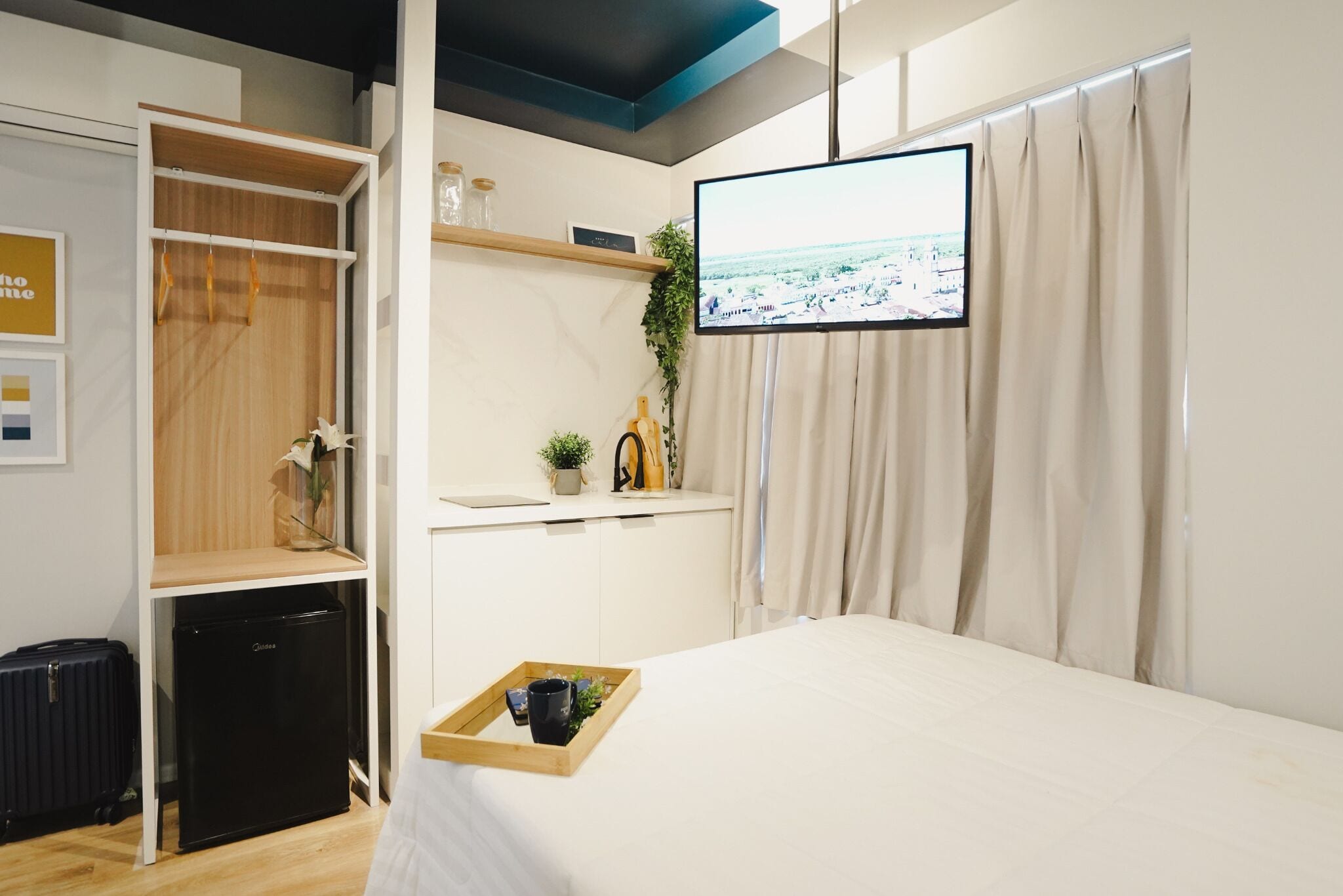 A hotel room with a bed, a TV mounted on the wall, a small desk with a plant, and a wardrobe with a hanging rack.
