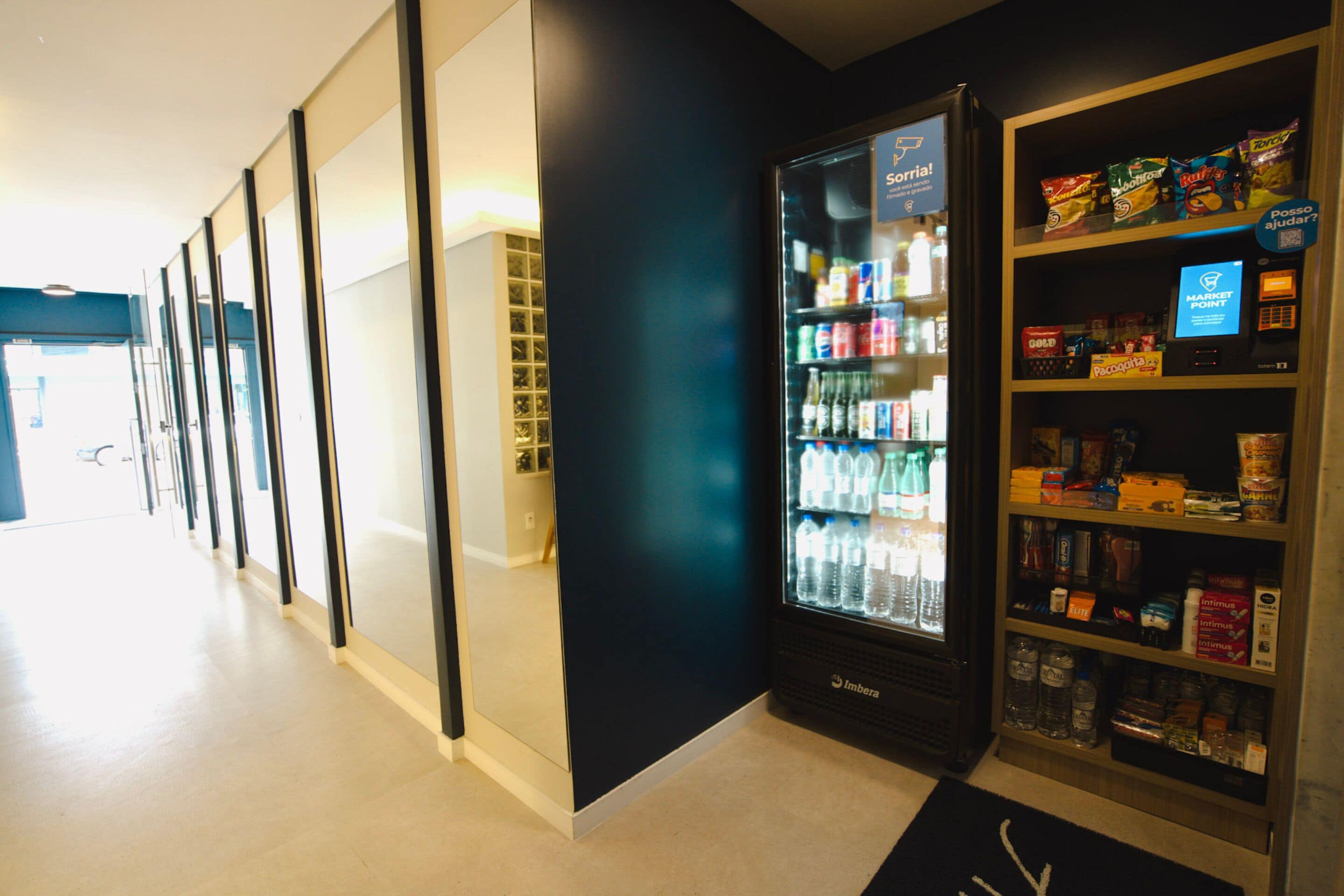 A vending machine with a variety of snacks and beverages, located in a hallway with glass doors and a black wall.