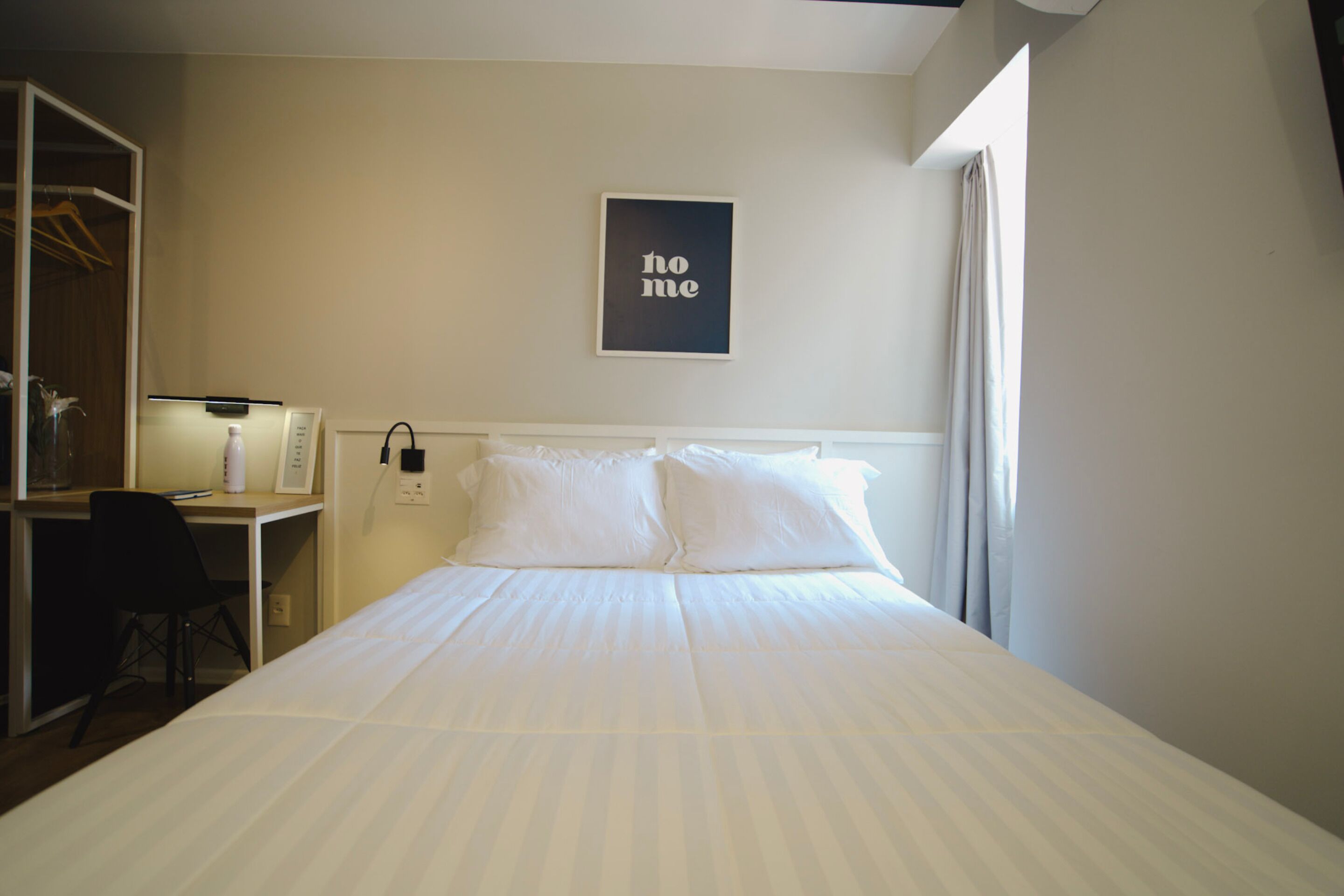 A hotel room with a bed, a desk, and a chair. There is a framed picture on the wall and a window with curtains.