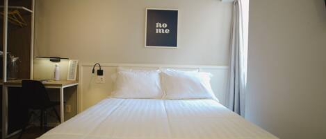 A hotel room with a bed, a desk, and a chair. There is a framed picture on the wall and a window with curtains.