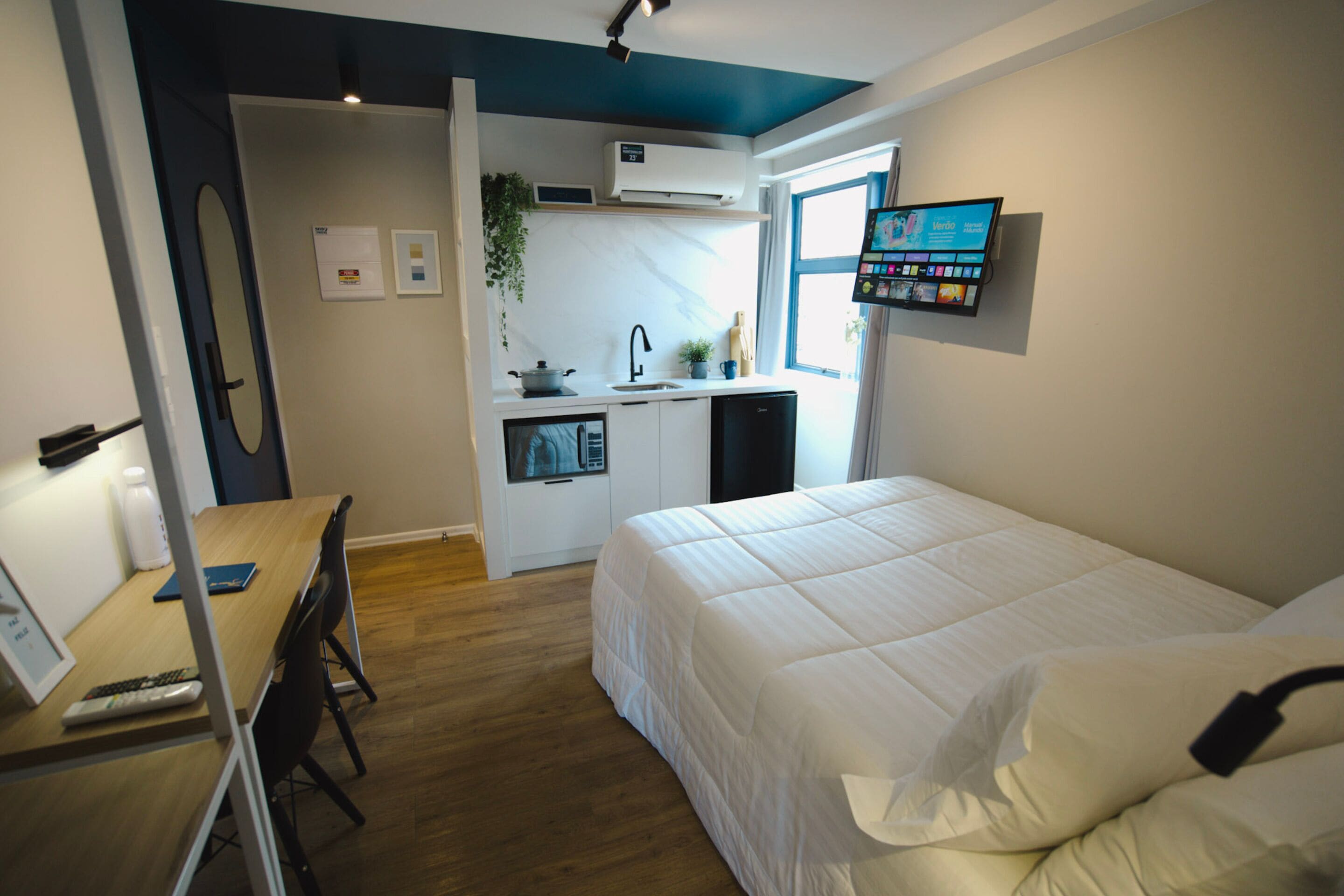 Habitación de hotel compacta con cama, escritorio con computadora, kitchenette pequeña y televisor montado en la pared.