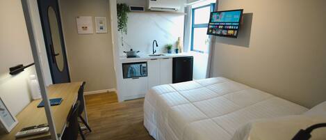 Habitación de hotel compacta con cama, escritorio con computadora, kitchenette pequeña y televisor montado en la pared.