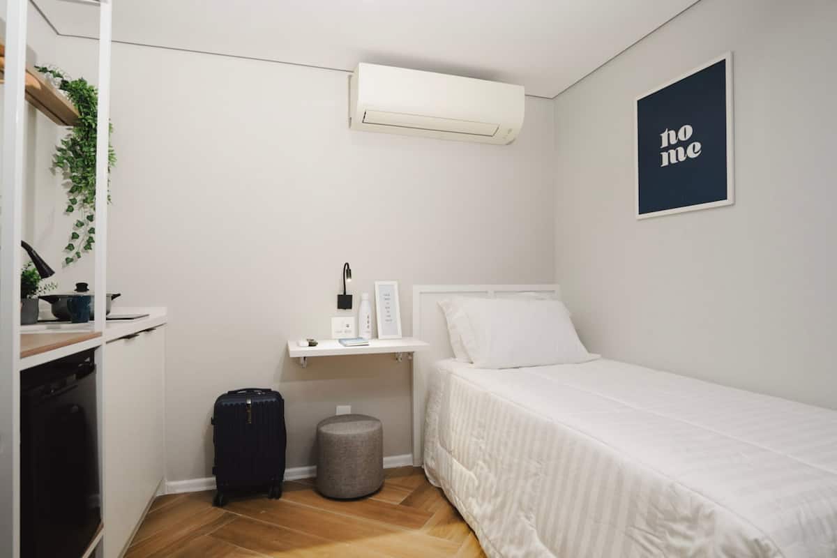 Une petite pièce, bien rangée, avec un lit, un petit bureau, un climatiseur fixé au mur, une poubelle et une pancarte indiquant « Ne pas déranger » sur le mur.