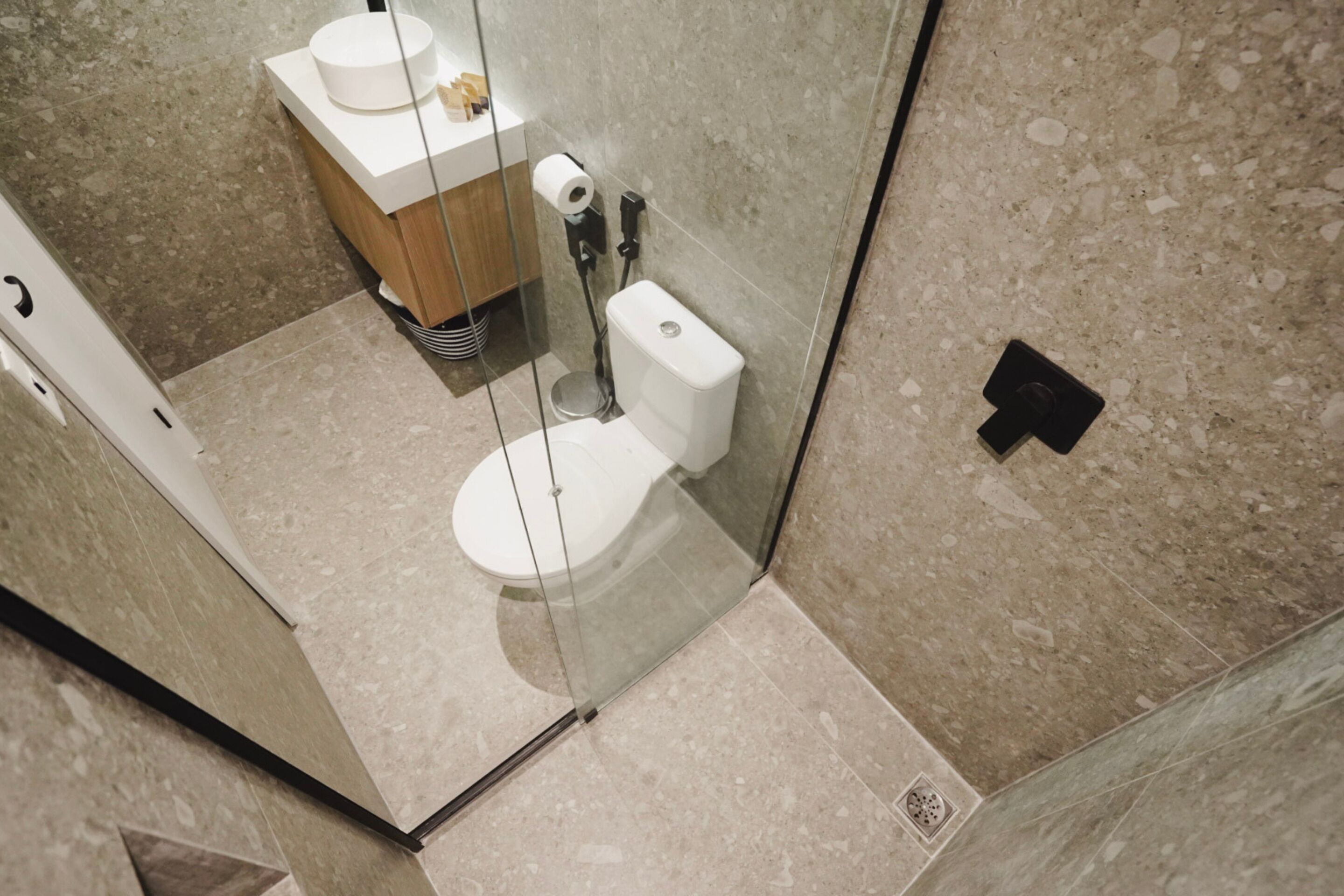 A modern bathroom with a glass-enclosed toilet, a wooden vanity, and a marble floor.