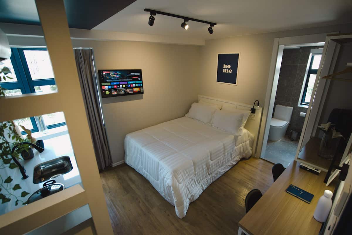 Une chambre à coucher avec un lit, un bureau, une chaise, une télévision et une salle de bain visible à travers une porte ouverte.