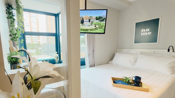 Habitación de hotel con cama, televisor mostrando vista a un jardín, un cartel que dice 'no me' y una bandeja con una taza y flores sobre la cama.