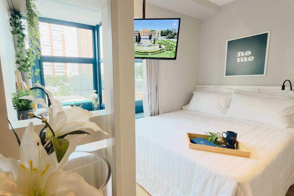 Une chambre d’hôtel avec un lit, un téléviseur diffusant une vue sur un jardin, une pancarte indiquant « Ne pas déranger » et un plateau avec une tasse et des fleurs posés sur le lit.