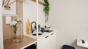 Una cocina compacta con un estante de madera, una planta colgante, un florero con una flor, una tetera y una taza azul.