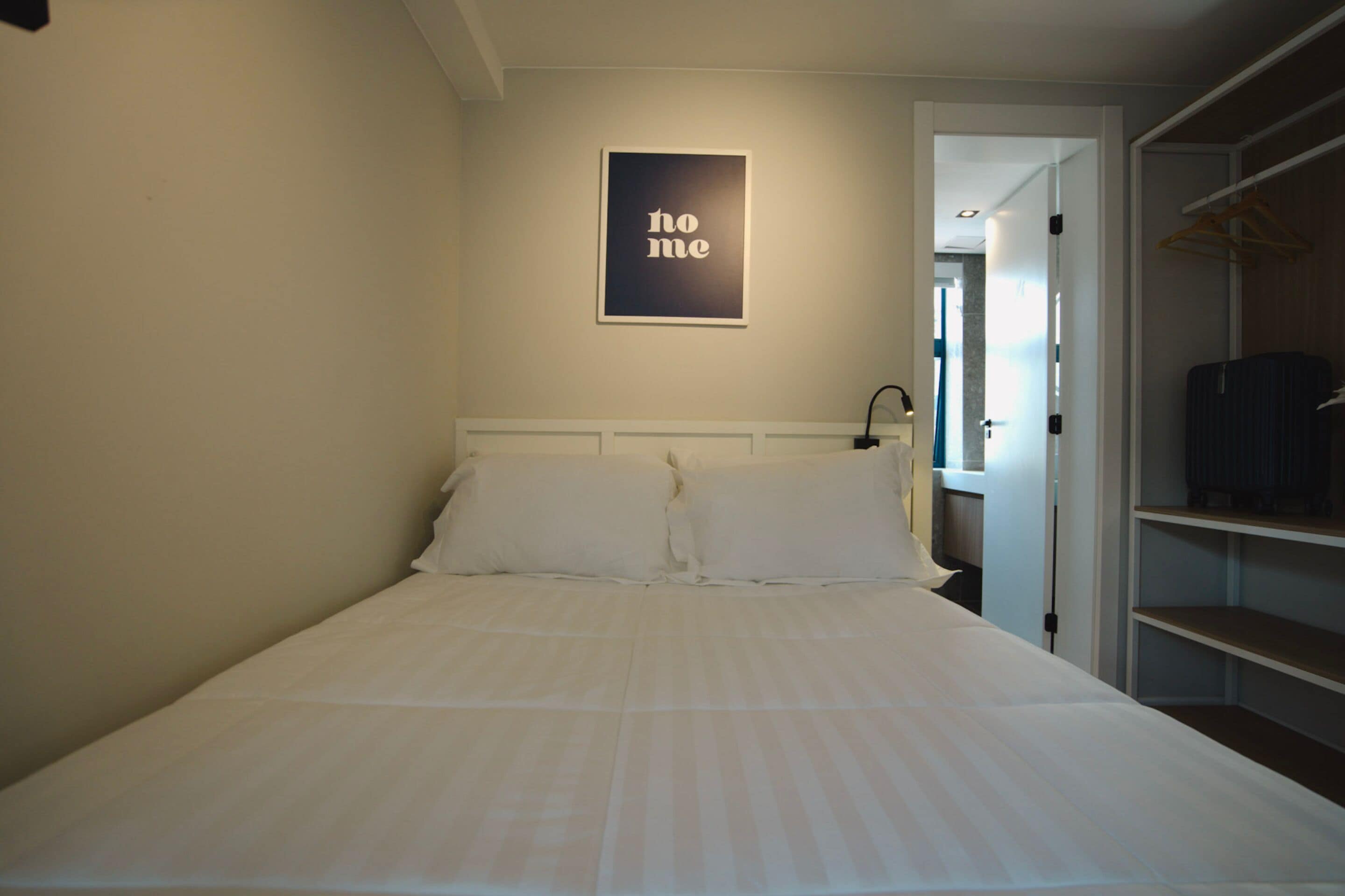 Ein ordentlich bezogenes Bett in einem Hotelzimmer, mit einem gerahmten Schild an der Wand und einem Kleiderschrank mit Kleidung.