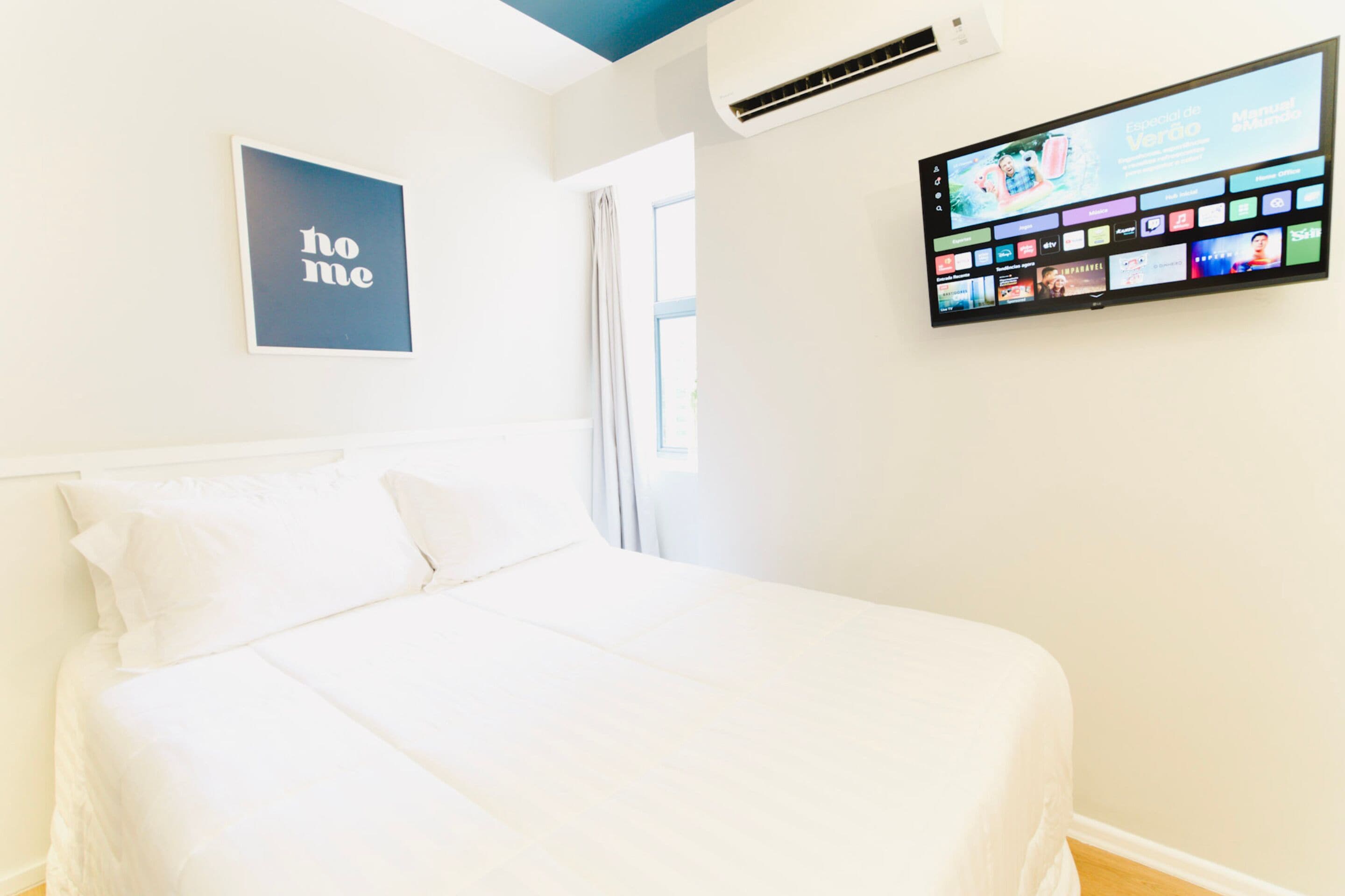 A hotel room with a bed, a 'home' sign, an air conditioning unit, and a digital screen displaying various channels.