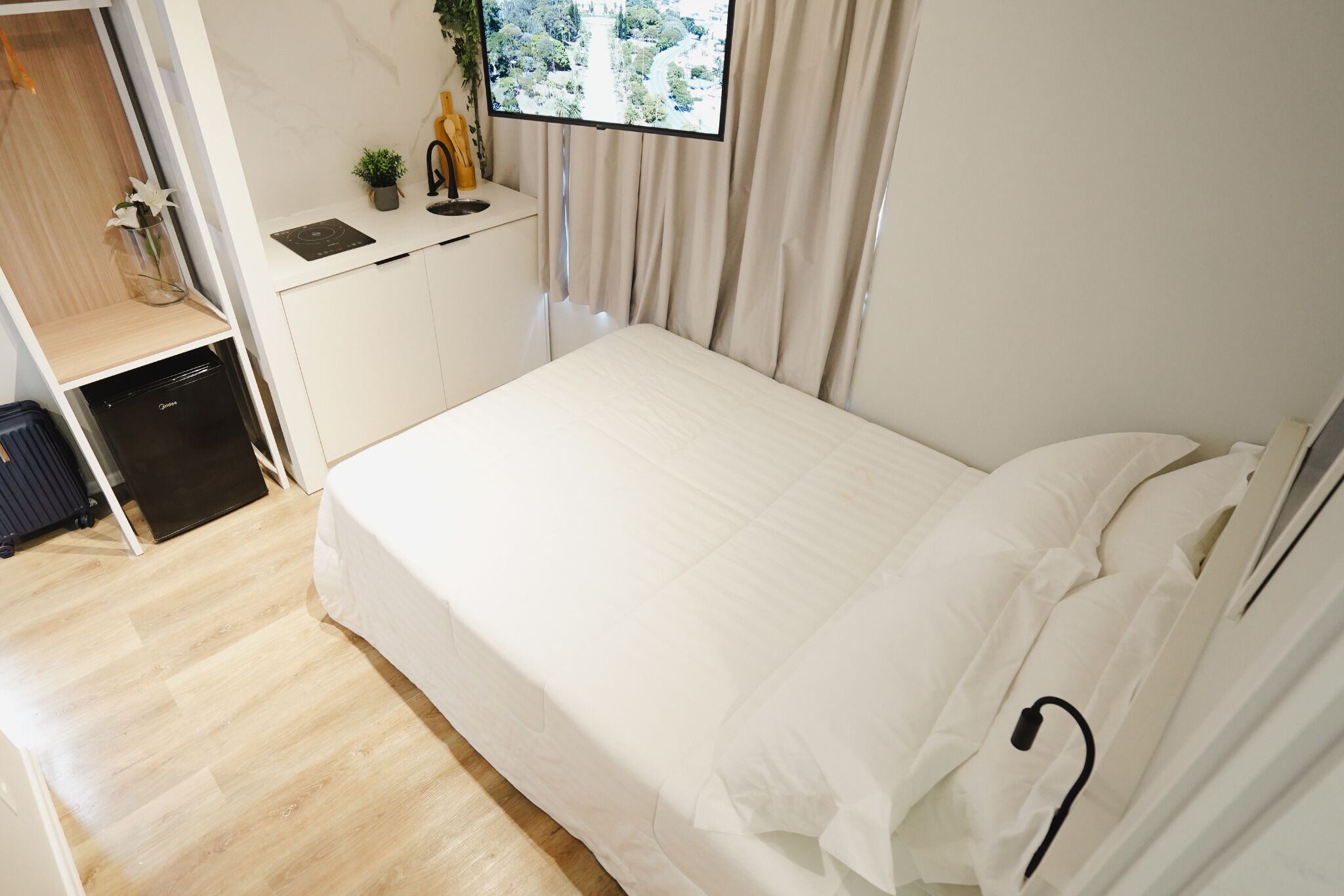 A neatly arranged bedroom with a bed, a small desk with a computer, a window with curtains, and a plant on the desk.