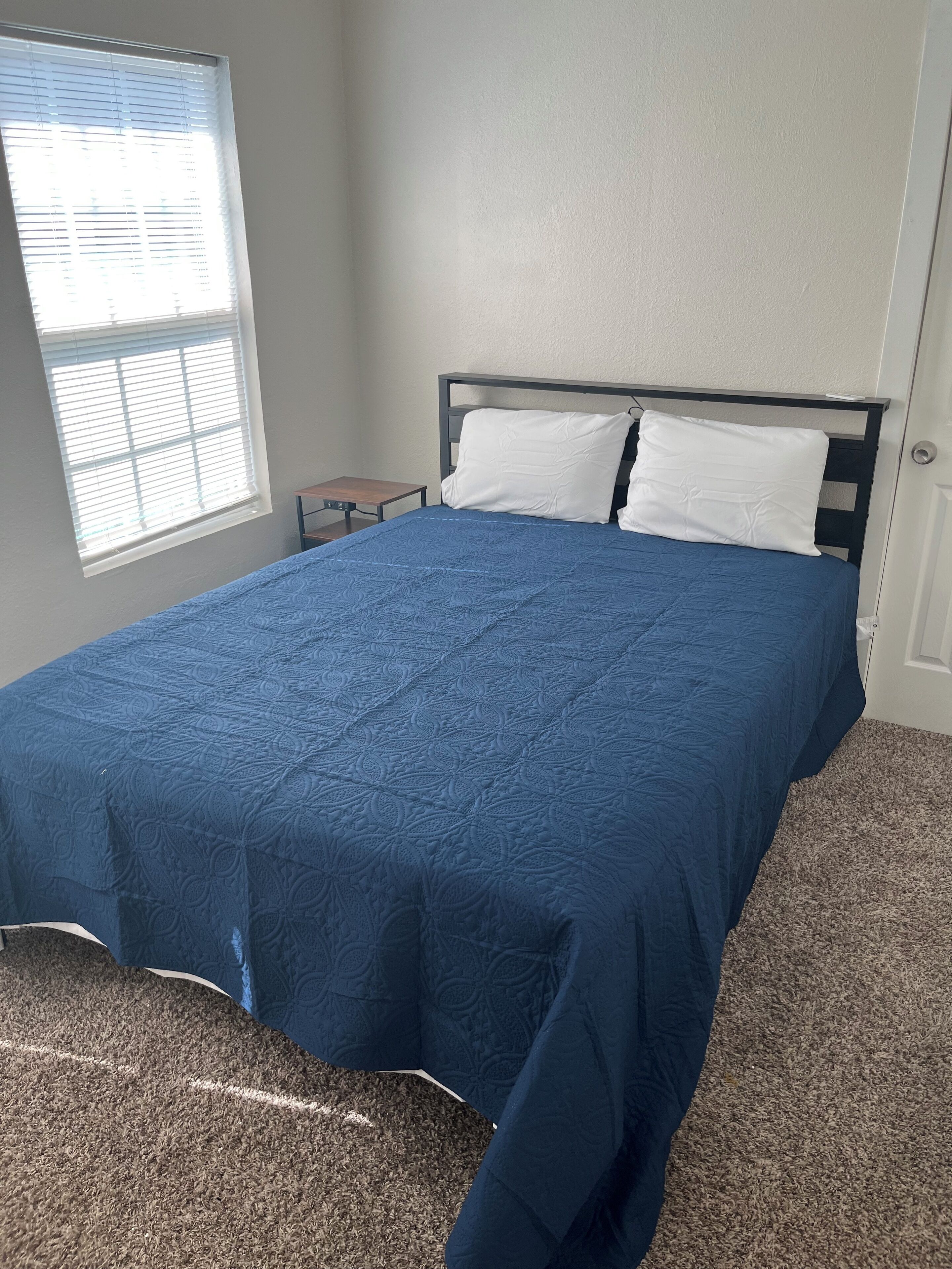 Un dormitorio con cama, ventana con persianas, una mesita y una puerta.