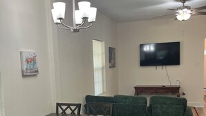 Un área de comedor con una mesa y sillas, un sofá verde, un televisor de pantalla plana y un ventilador de techo.