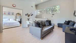 A modern living room with a grey sofa, a grey armchair, and a black coffee table. There is a white desk with a chair, a small plant, and a clock on the wall. The room has large windows and a ceiling fan. - 1 BR Guest House, Full Kitchen, 3-minutes to Downtown Austin! (Austin)