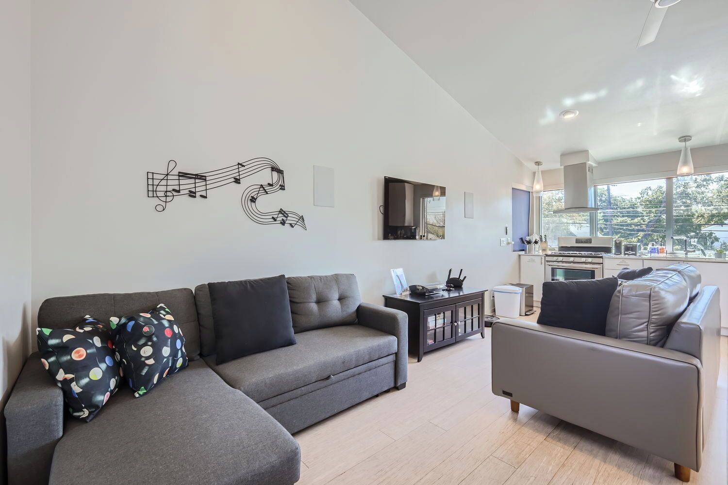 A modern living room with a grey sofa, a grey armchair, and a coffee table. There is a kitchen area with a stove and a window overlooking a garden.