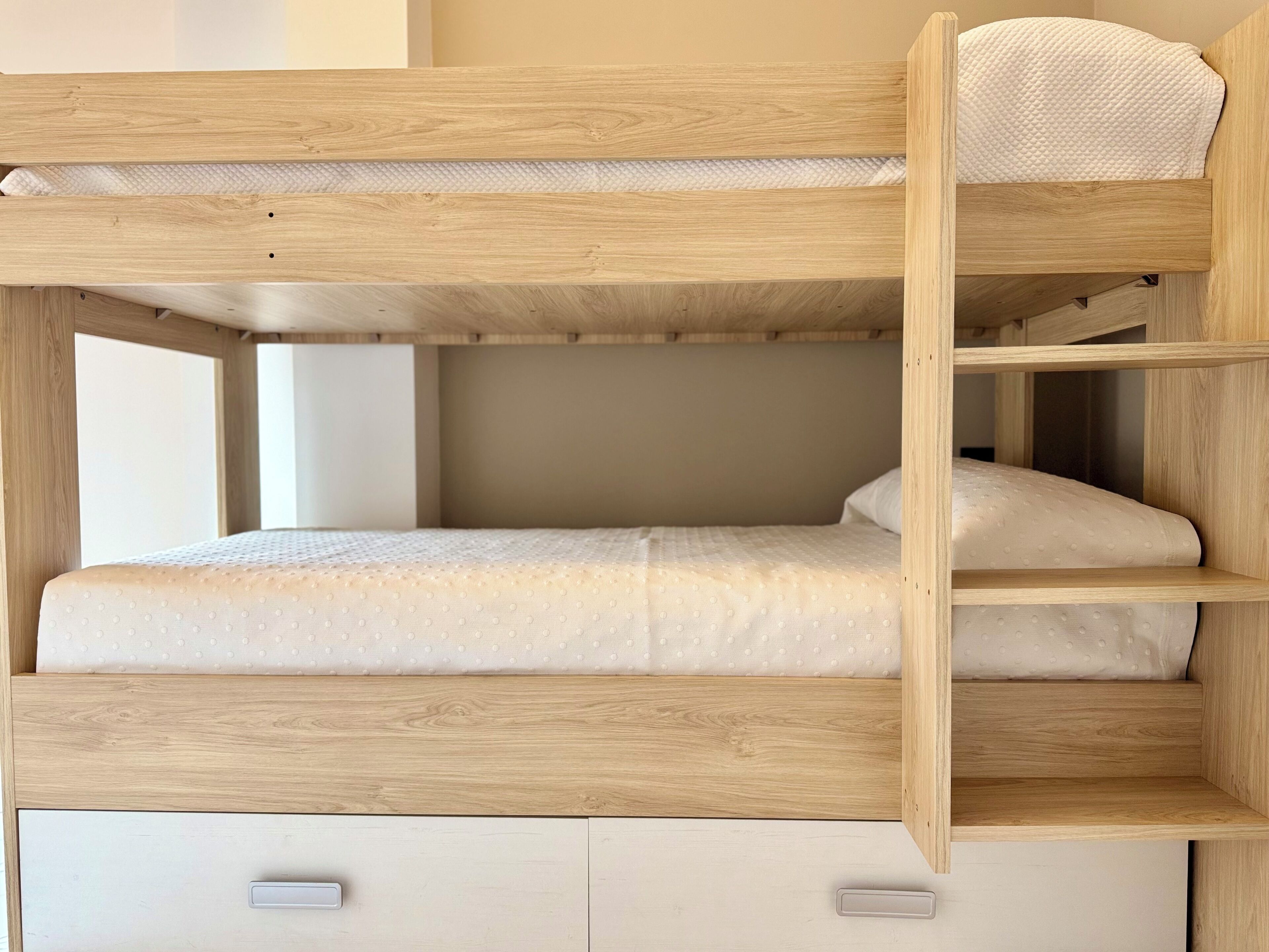 Una cama litera con estructura de madera, ropa de cama blanca y un estante pequeño.