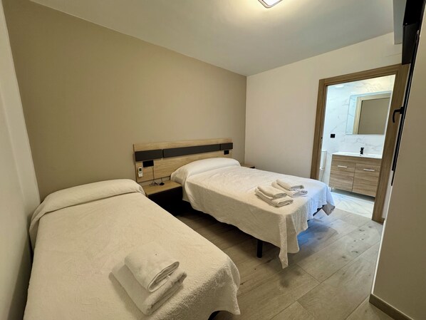 Ein Hotelzimmer mit zwei Betten, einem hölzernen Kopfteil, einem kleinen Nachttisch mit Telefon sowie einem durchgehenden Türspalt einsehbaren Badezimmer.