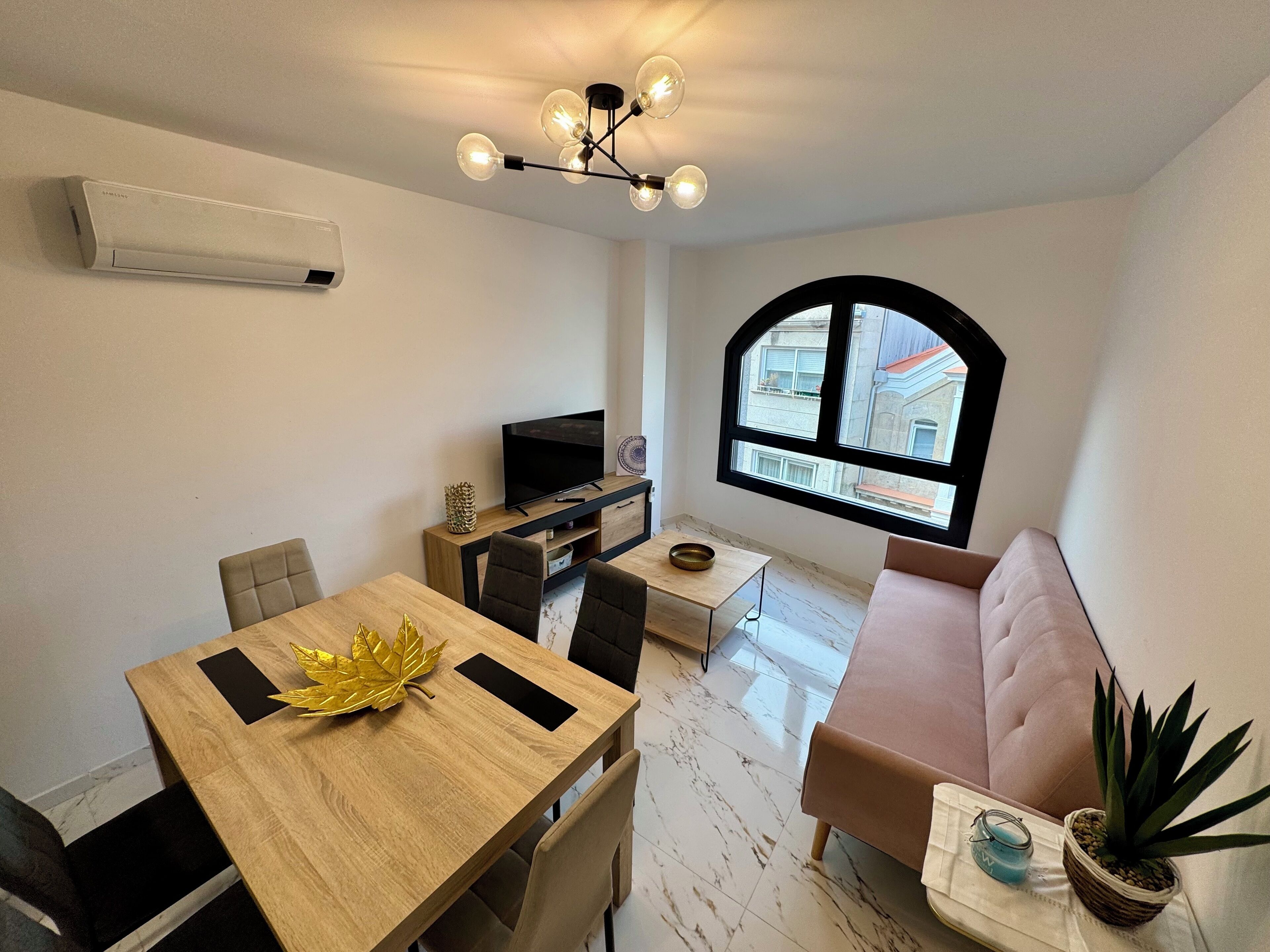 Una sala de estar moderna con una mesa de comedor de madera, un sofá, un televisor y un ventanal con vistas a edificios.