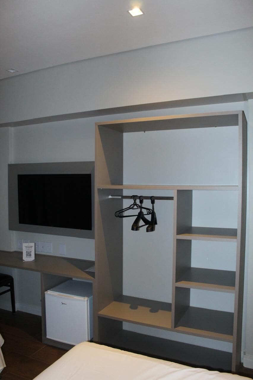 A room with a bed, a TV mounted on the wall, a small fridge, and a wardrobe with shelves and hanging space.