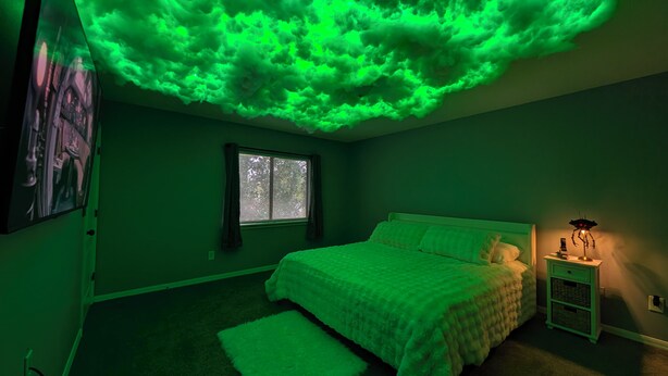 Un dormitorio con cama, ventana, mesita de noche y un proyector de pared que muestra una proyección de nube verde.