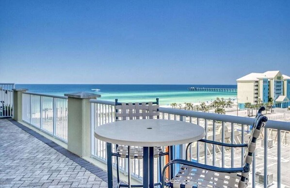 A balcony with a round table and chairs, overlooking a beach with a pier in the distance.