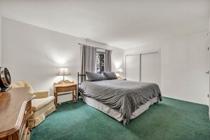 Ein Schlafzimmer mit einem Bett, einem Schreibtisch, einem Sessel, zwei Nachttischlampen, einem Fenster mit Vorhängen und einer Tür.
