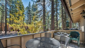 Eine überdachte Terrasse im Freien mit einem runden Metalltisch und Stühlen, umgeben von hohen Bäumen und blauem Himmel.