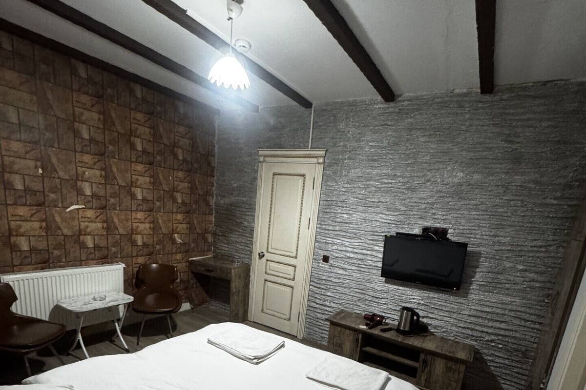 Un dormitorio con cama, una silla, una mesita, un radiador, una puerta, un televisor y un banco de madera.