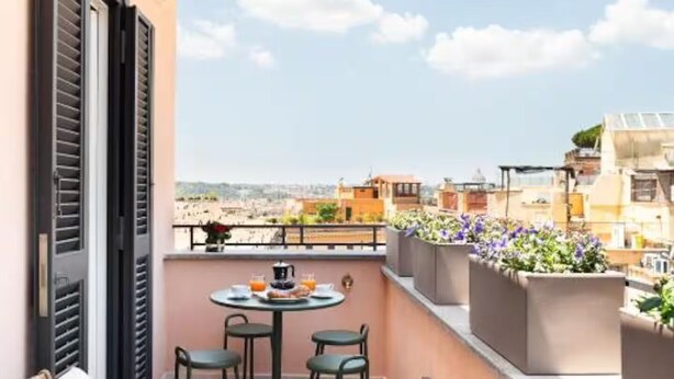 A balcony with a white sofa, two chairs, a small round table with a coffee pot, and a view of the cityscape.
