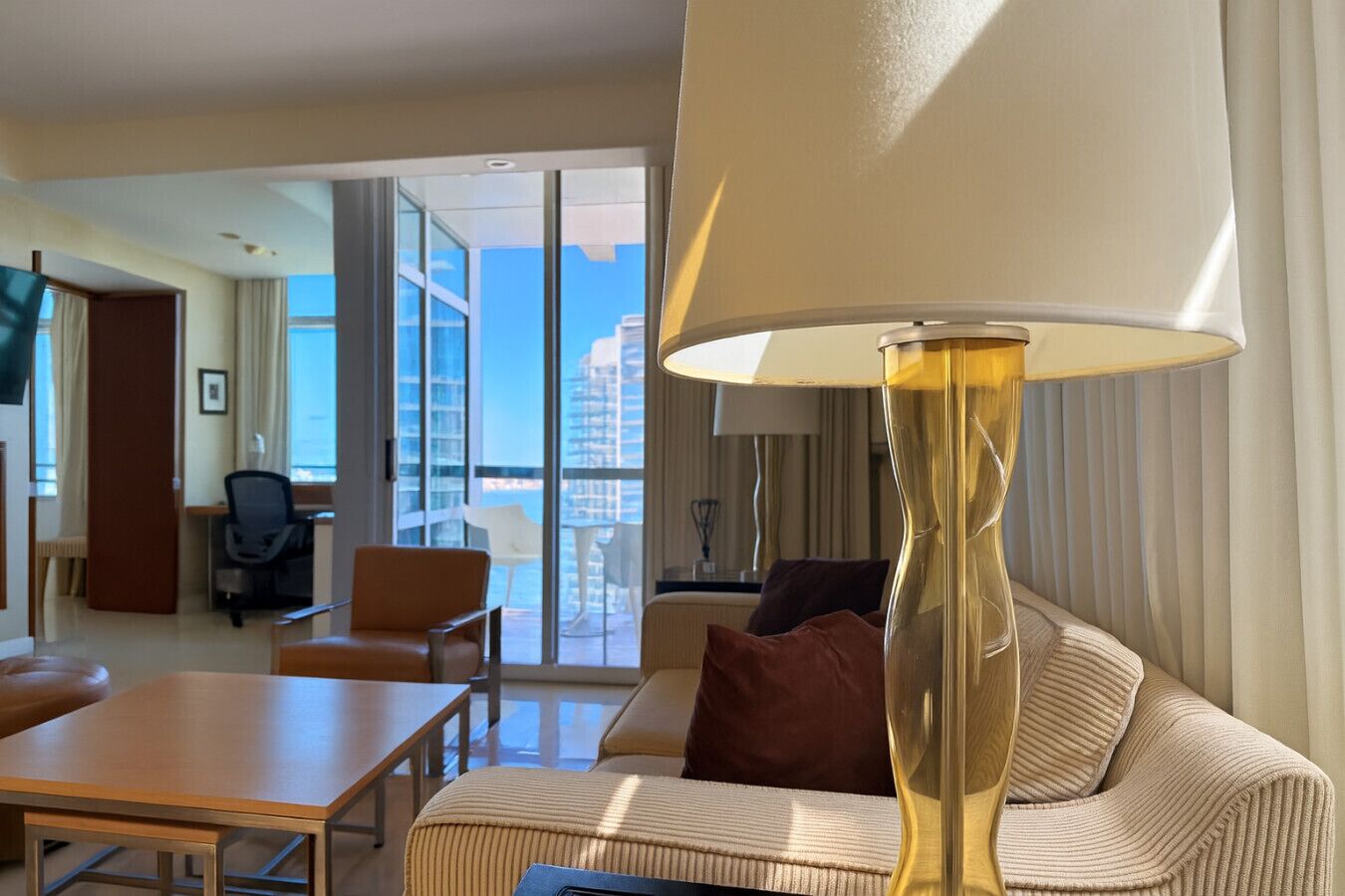 Ett hotellrum med en soffa, ett bord, en lampa och utsikt över staden genom stora fönster.