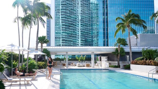 A pool area with lounge chairs and umbrellas, surrounded by palm trees and a modern building with reflective glass windows. - AKA Hotel Stunning Views w- Free parking (Miami)