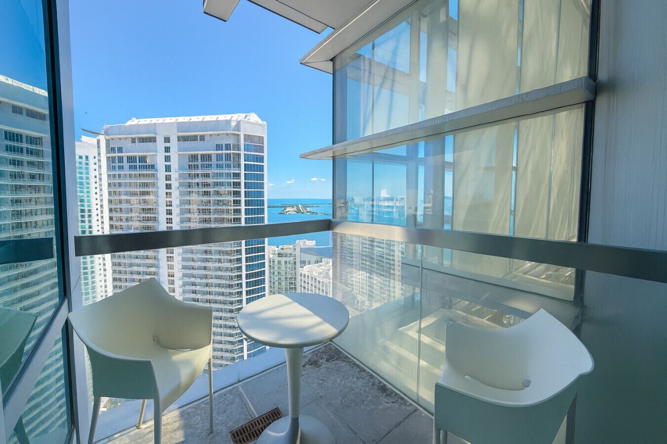 A balcony with two white chairs and a small table, overlooking a cityscape with tall buildings and a view of the ocean.