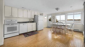 A kitchen with white cabinets, a stove, and a refrigerator. There is a dining table with chairs and a view of the water through large windows. - Cozy Lakefront 2bd Duplex on Long Lake (Traverse City)