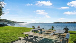 A lakeside picnic area with picnic tables and chairs, a small dock, and a grassy lawn. - Cozy Lakefront 2bd Duplex on Long Lake (Traverse City)