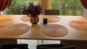 Een eettafel met uitzicht op de zee door grote ramen, bruine gordijnen en een potplant.
