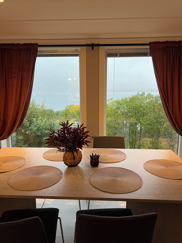 A dining table with a view of the sea through large windows, brown curtains, and a potted plant.